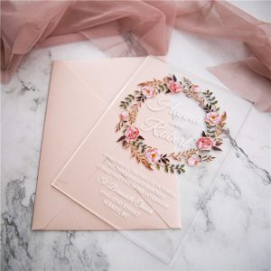 Cette photo représente un faire part de mariage imprimé sur du plexiglass avec son enveloppe assortie de couleur rose poudré. Le motif du faire part est articulé autour d'une couronne fleurie dans les tons de roses et de verts